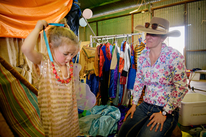 A little girl putting a necklace over her head as a woman wearing a cowboy hat watches on, inside a large shed. 